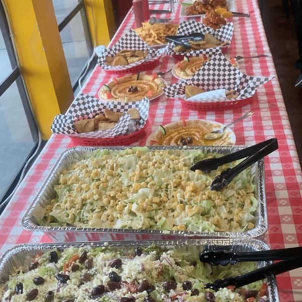 A buffet table with salads, hummus, pita bread, and french fries on a red and white checkered tablecloth.